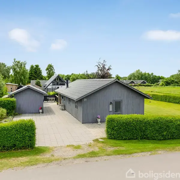 Et gråt enfamiliehus med et skur ved siden af hviler stille på en flisebelagt indkørsel. Omkring huset er en velplejet græsplæne og grønne buske i en landlig, grøn omgivelser.