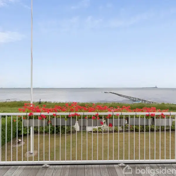 Terrasse med hvidt gelænder og røde blomsterkasser, der vender ud mod en stor have. I baggrunden ses stranden og havet med en lang badebro strækkende ud i vandet.