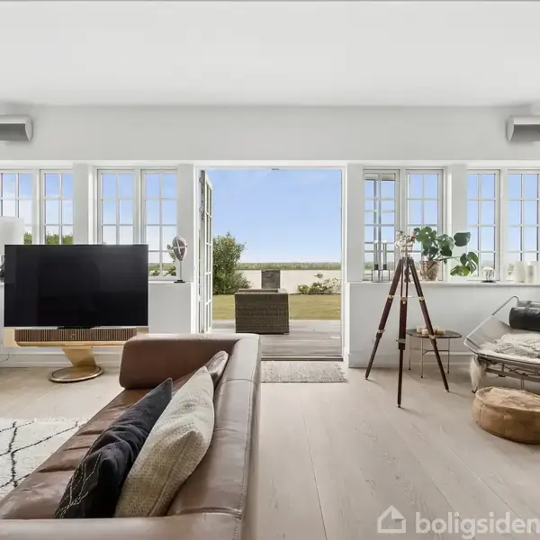 Stue med sofa og fladskærms-TV, der står foran store vinduer. Til højre ses en lænestol, et lille bord og en kikkertstativ, med udsigt til en have gennem åbne døre.