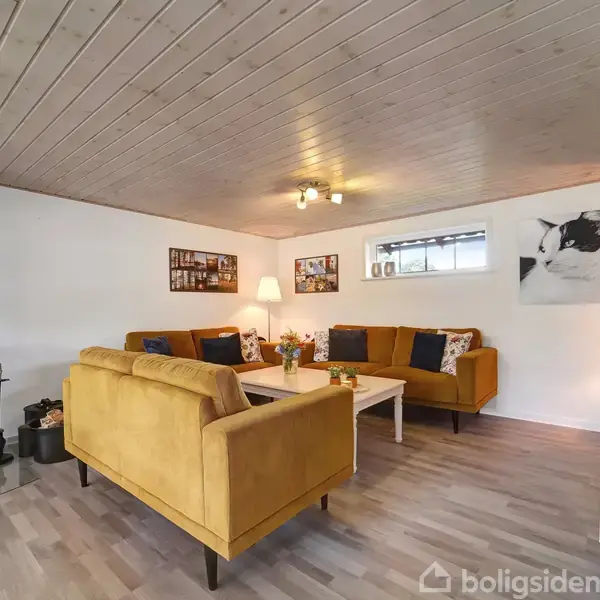 Brændeovn brænder i stue med lyst træloft. Gyldne sofaer omkranser et hvidt sofabord med blomster. Billeder hænger på væggene, og et lille vindue slipper lys ind.