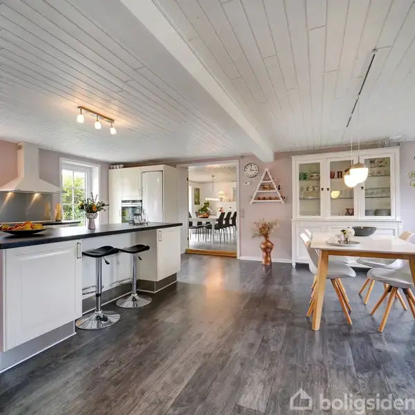 Moderne køkkenalrum i åbent layout. Et køkkenbord med barstole står til venstre, mens et spisebord med fire stole er til højre. Store vinduer og planter skaber en lys atmosfære.