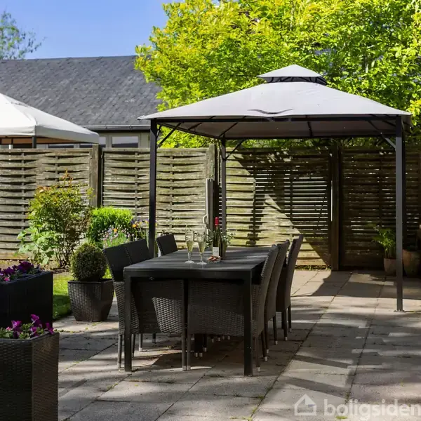 Et udendørs spisebord med seks stole står under en pavillon i en flisebelagt gårdhave. Omgivet af blomster og hække, med et plankeværk og naboens tag i baggrunden.