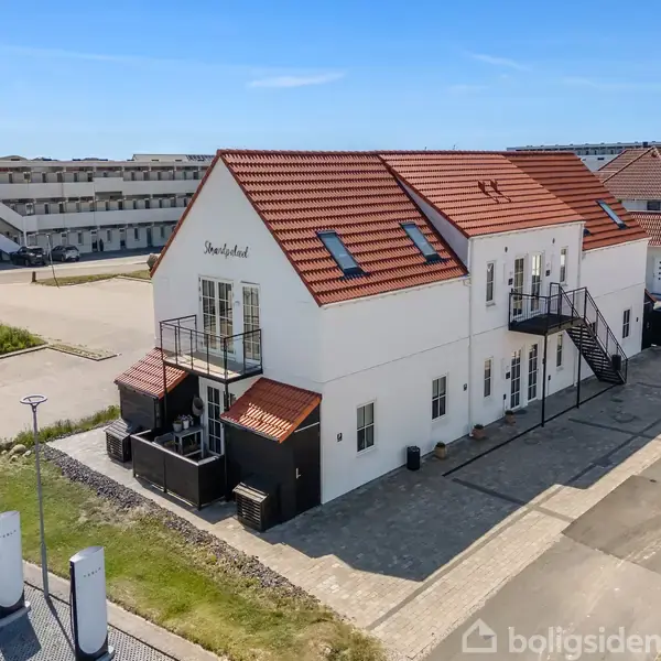 Hvid bygning med rødt tegltag og sorte trapper, står på asfalteret område omgivet af parkering og andre bygninger. Tekst på facaden: "Strandhotel."