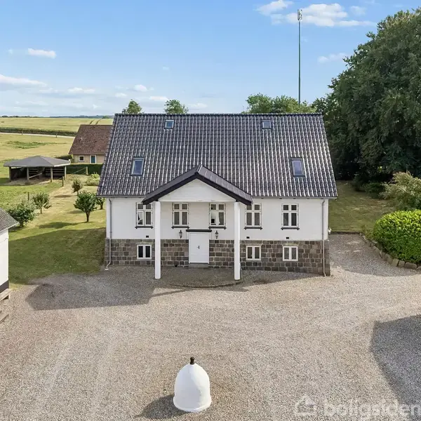 Bygning står stille på en gård omgivet af grusplads og grønne træer. Huset er hvidt med gråt tag, flankeret af to mindre bygninger på hver side.