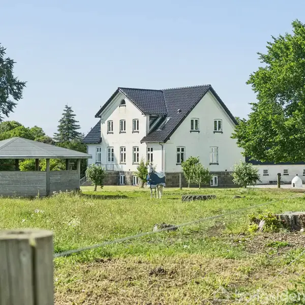 Hvidt hus står stille på en stor grøn eng med et lyst træpavillon i nærheden. Træer omkranser området, der er indhegnet med lave pæle.