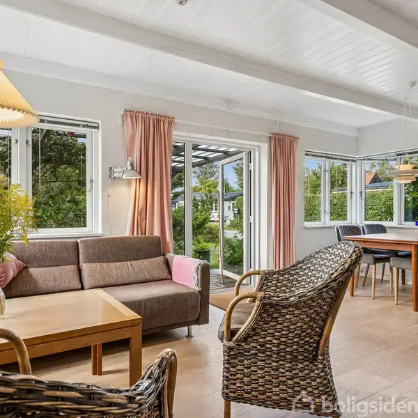 Stue med sofa og kurvestole omkring et sofabord; store vinduer giver udsigt til en grøn have. I hjørnet står et spisebord med stole. Lampelys tilfører hygge.