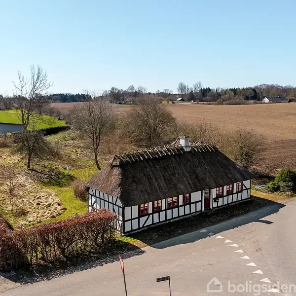 Stråtækt bindingsværkshus står stille ved en vej i et landligt område med åbne marker og spredte træer. Husets facader er sort-hvide med røde vinduesrammer.