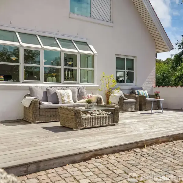 Terrassemøbler står på en træterrasse ved et hvidt hus med store vinduer. Møblerne omfatter en sofa, lænestol og sofabord med puder og planter, omgivet af grønt område og brosten.