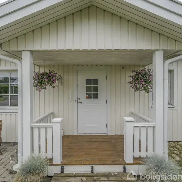 Husets indgang har en hvid dør med små ruder, hængende blomsterkurve på hver side. Til venstre står et sæt havemøbler på en stenbelagt gårdsplads, omgivet af græsplæne.