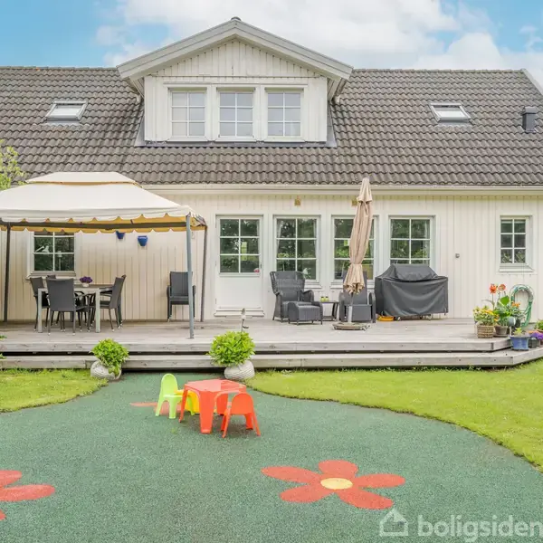 Et hvidt hus har en terrasse med parasol, borde og stole i en have. Foran terrassen er der et legeområde med børnemøbler og farverige blomsterdekorationer på jorden.