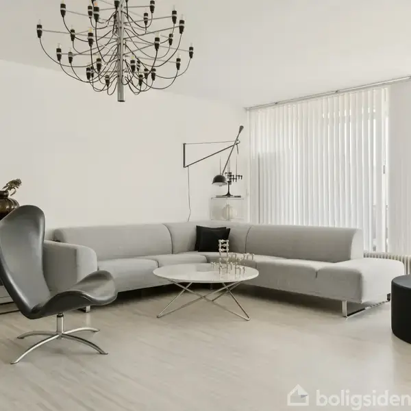 En moderne stue med en stor, grå hjørnesofa står ved siden af en kurvet sort lænestol. En rund, hvid kaffebord og en stor lysekrone hænger fra loftet. Tykke gardiner dækker vinduerne.