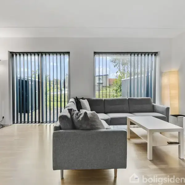 Grå sofa står i stue ved to store vinduer med persienner, omgivet af en lampe og et sofabord, mens et fjernsyn er placeret til højre.