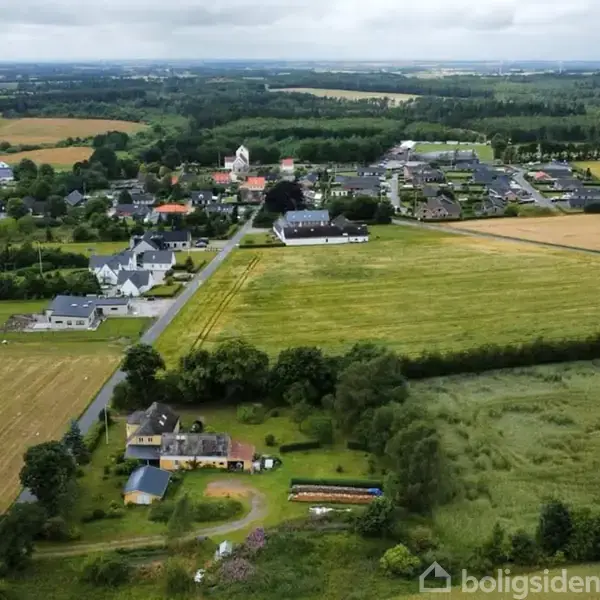 Rødt hus ligger i landsby omgivet af marker. En rød pil peger mod mark til venstre. Området er præget af grønne arealer og forskellige bygninger i baggrunden.