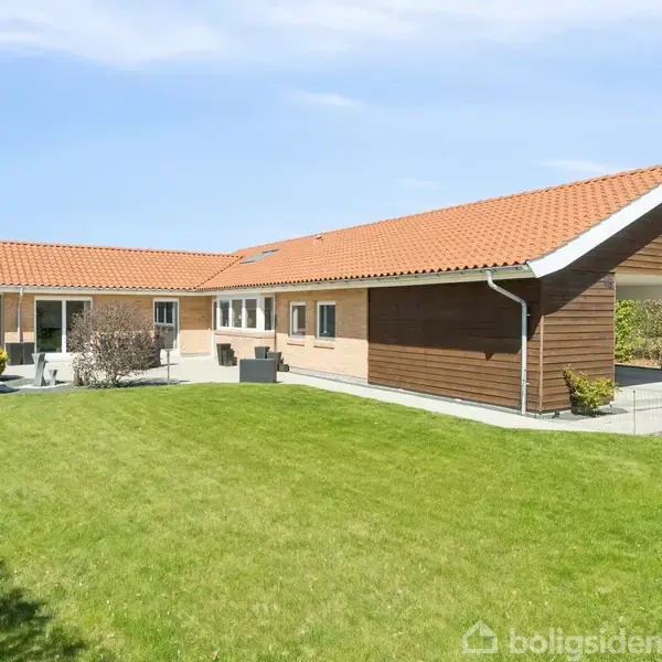Et moderne, enfamiliehus med rødt tegltag står på en velplejet græsplæne. Huset har store vinduer, og i baggrunden ses en blå himmel og nogle buske langs hegnet.