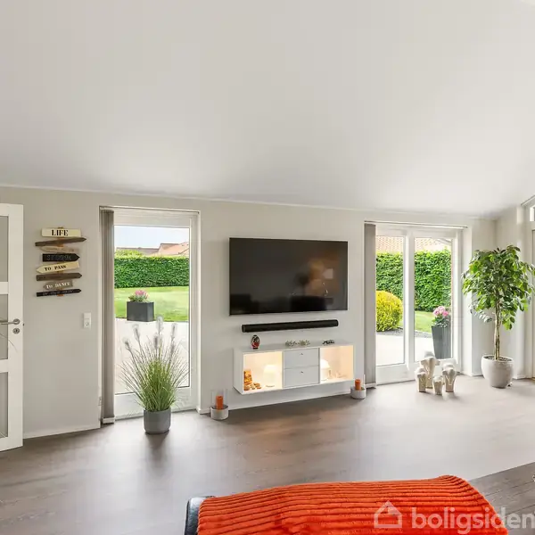 Et moderne hus med rødt tegltag står blandt velplejede grønne haver og hække. En flisebelagt indkørsel fører op til hoveddøren. Omgivelserne er et grønt boligområde.