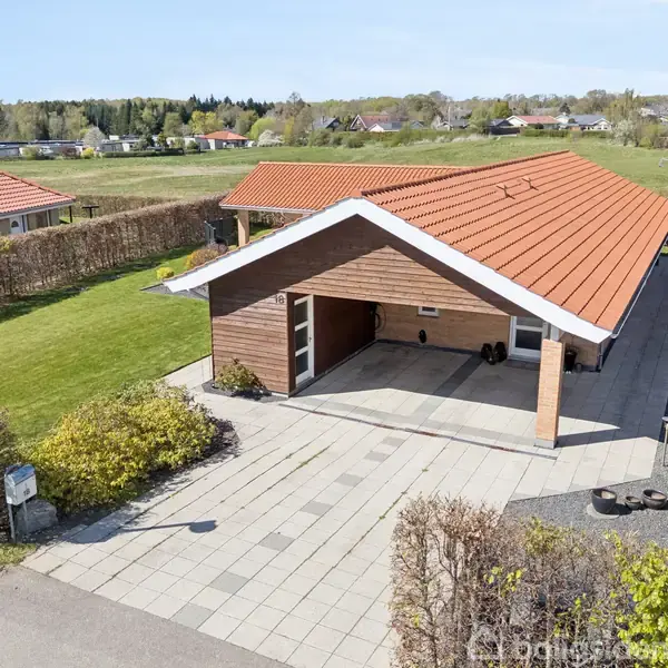 Et moderne hus med rødt tag står stille i en velplejet have med grønne buske og en flisebelagt indkørsel. Omgivet af hække i et villakvarter.