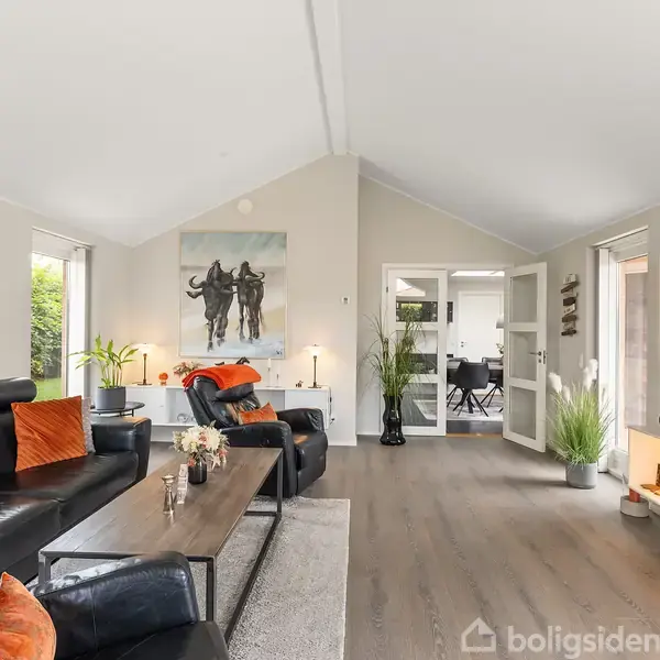 Et moderne hus med rødt tegltag og en carport står i en velplejet have omgivet af hæk. I baggrunden ses et grønt område med flere huse.
