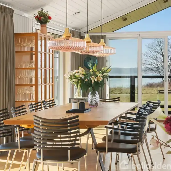 Et spisebord med sorte stole står i et lyst rum med store vinduer, der viser udsigt til have og vand. Over bordet hænger to lysekroner. Der er blomster på bordet.