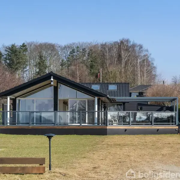Et moderne sommerhus med glasfacade står stille på en plæne omgivet af grønne buske og træer i baggrunden, under en klar blå himmel.
