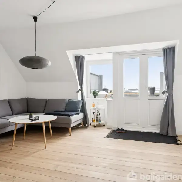 Grå hjørnesofa står mod væggen i en stue med trægulv. Et rundt bord står foran sofaen, og der hænger en pendellampe over bordet. Døre fører ud til altan. Enkelt dekoration i rummet.