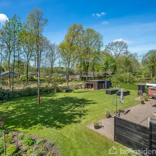 En stor, grøn græsplæne med flagstang og terrasse tilknyttet et hus. Der er træer i baggrunden, der danner et frodigt skovområde under en klar blå himmel.