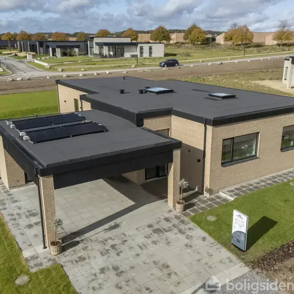 En moderne, fladtagsvilla i mursten står på et nyanlagt boligkvarter med grønne plæner og små træer. Foran huset er en indkørsel. En skilt med tekst står ved indgangen.