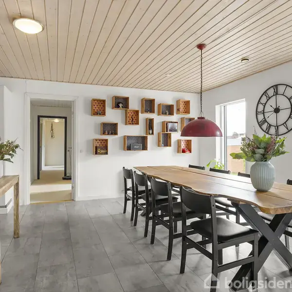 Langt spisebord med otte stole står i et lyst, moderne spisestue. Væggen har dekorative hylder, og en stor, rund ur hænger ved siden af to store vinduer.