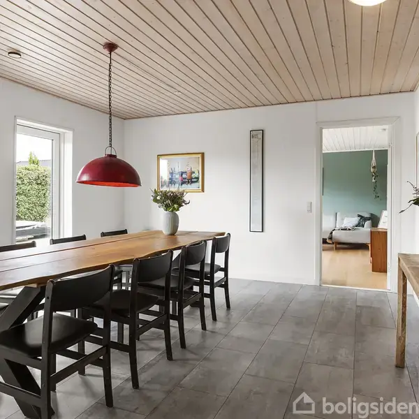 Et langt træbord med sorte stole står i en spisestue. En rød lampe hænger over bordet, og der er en stor, rund vægur på væggen. En åben dør fører til stuen.