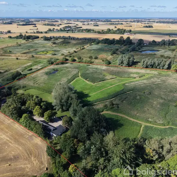 Hus omgivet af træer og marker, beliggende i et stort landligt område med marker og skove. En sti slynger sig gennem det grønne landskab, omgivet af blå himmel og få skyer.