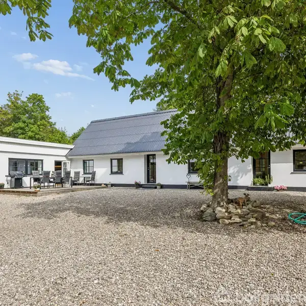 Et hvidt hus med sort tag står omgivet af grus, træer og græs. En terrasse på siden af huset rummer havemøbler under en klar blå himmel.