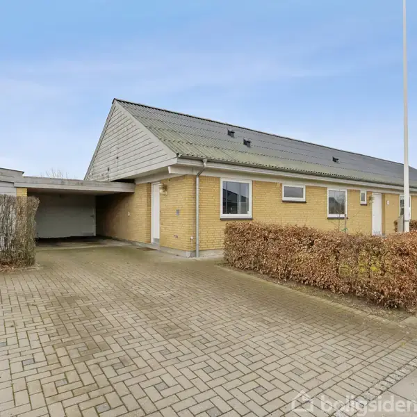 Hus i mursten ved siden af en garageplads. Bygningen er omkranset af en hæk, og belægningen er af betonfliser. Baggrunden viser en klar, blå himmel.