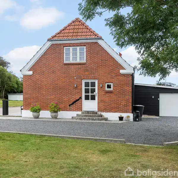 Rødt murstenshus med skråt tag, stående på grusplads. Nogle blomsterkrukker står foran indgangen. Omkring er der græsplæner og træer set under en delvist skyet himmel.