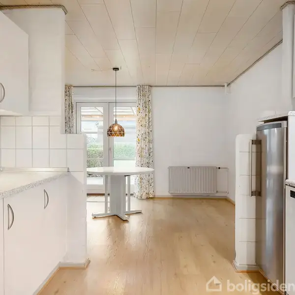 Køkken med hvide skabe og fliser i handlingen med at omkranse rummet. Trægulv og spiseplads i kontekst med vindue og hængende lampe i baggrunden.