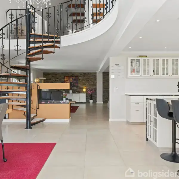 En spiraltrappe af træ og metal fører op til en balkon i en stilren stue med lys indretning. Der er et køkken med barstole og lyse klinkegulve.