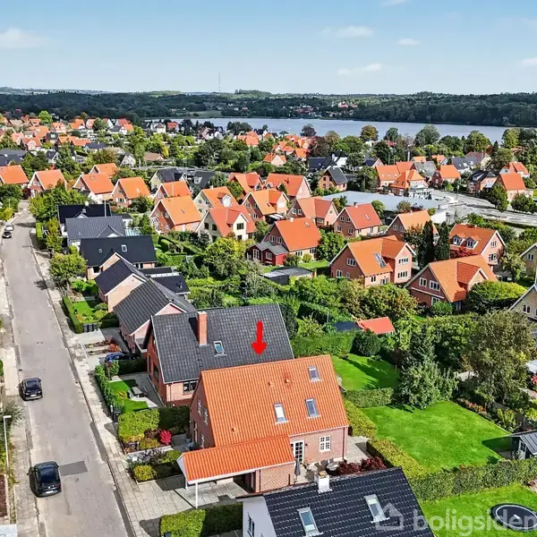 Rødt teglhus markeret med pil; står stille i villakvarter med mange lignende huse, græsplæner og haver. Set fra oven med udsigt over sø i baggrunden.