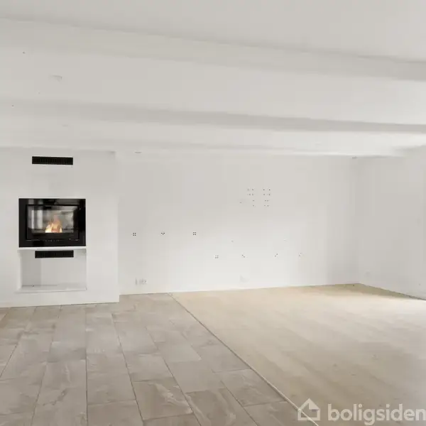 Stue med pejs, delvist dækket af flise- og trægulv, indbygget i en hvid væg. Store vinduer på højre side slipper lys ind i rummet.