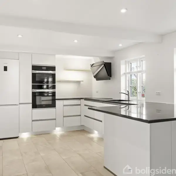 Et moderne køkken med hvide skabe og sort bordplade. Køkkenet har indbygget ovn, komfur og emhætte. Stort køleskab til venstre. Rummet er lyst med store vinduer.