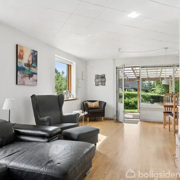 Sort lædersofa står i en moderne stue med lyse vægge og trægulv. Stolen vender mod et stort vindue med udsigt til en grøn have. Rummeligt og lyst interiør.