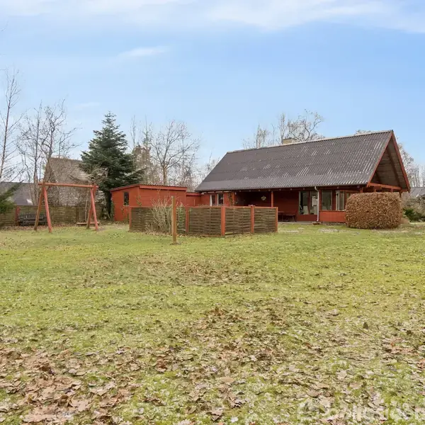 Rødt træhus med skråt tag står i en stor have, omkranset af træer. En hvid bil ses til højre, og en gyngestativ er placeret til venstre.