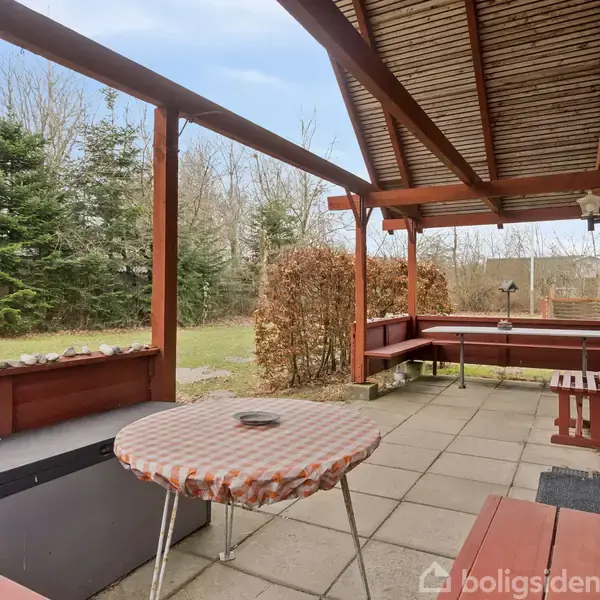 Et overdækket terrasseområde indeholder et bord med ternet dug, en bænk og træstole. Terrassen er omkranset af et hegn og ligger i en have med græs og træer.