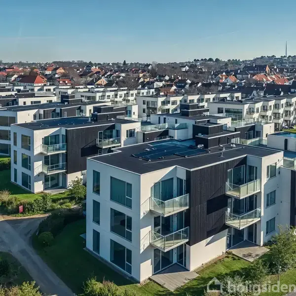 Boligkomplekset består af moderne lejlighedsbygninger med altaner, omgivet af grønne områder og stier. I baggrunden ses en by med mange tage og spredte bygninger.