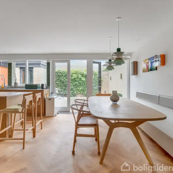 Et moderne køkken-alrum med trægulv, hvor et træbord står centralt omgivet af stole. Panelet til højre har indbygget siddeplads. Store glasdøre fører ud til en grøn hæk.