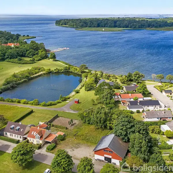 Boliger ligger ved en sø nær kysten, omgivet af grønt landskab og træer. En mindre vej snor sig gennem området, der giver adgang til husene.