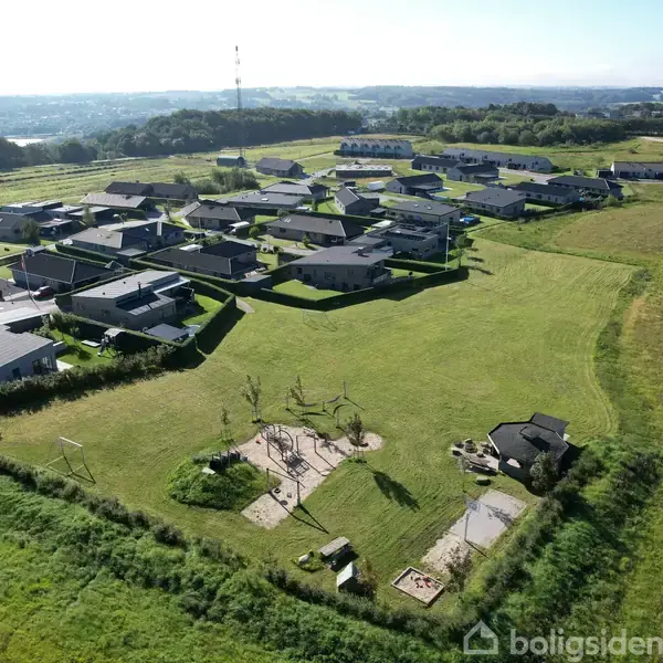 En række moderne huse ligger i en ny boligudvikling omgivet af store grønne marker. En legeplads med gynger og sandkasse ses i forgrunden.
