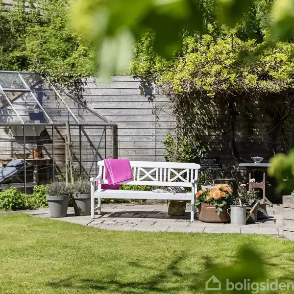 En hvid bænk med en pink pude står på en terrasse foran et drivhus i en frodig have. Planter og blomster i potter omgiver bænken, mens grønne træer omkranser området.