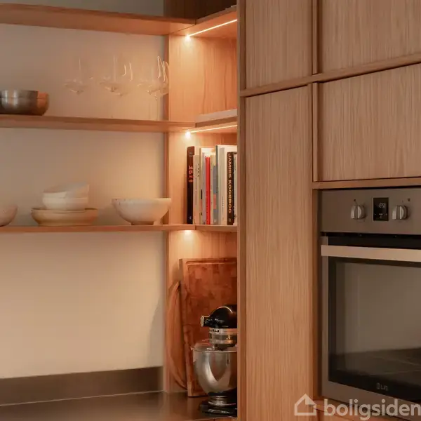 Træhylder fyldt med skåle, glas og bøger er placeret ved siden af en ovn. Miljøet er køkkenet, og blødt indbygget lys lyser op på hylderne.