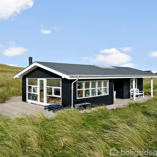 Sort sommerhus står stille blandt lange græsstrå. Store vinduer og terrassemøbler pryder ydersiden. Omgivet af åbne græsarealer under en blå himmel med få skyer.