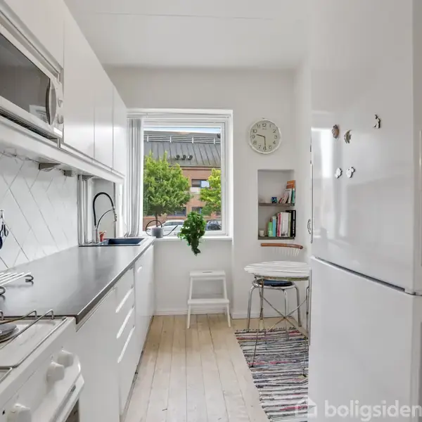 Et smalt køkken med hvide skabe og apparater. Stolen står foran et vindue, hvor en grøn plante er synlig. En hylde indeholder bøger, og et ur hænger på væggen.