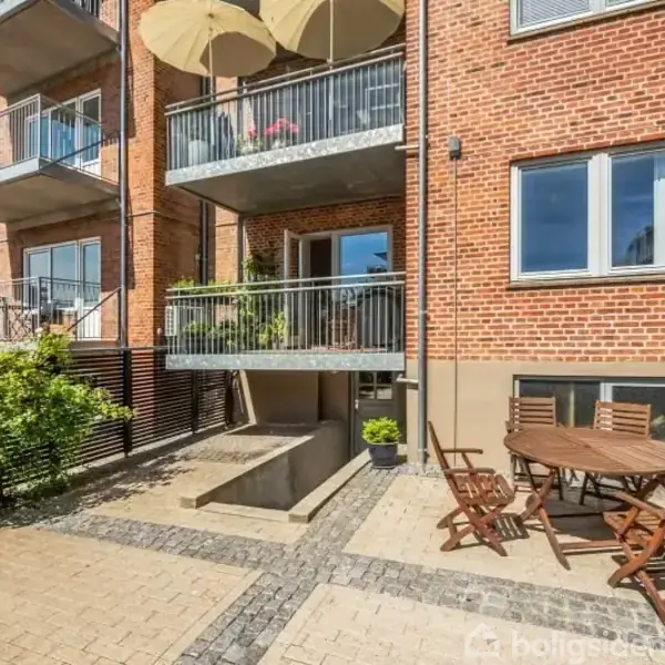 Udendørs terrasse med rundt bord og fire stole placeret på flisebelagt område, omgivet af cykler og en murstensbygning med altaner i baggrunden.