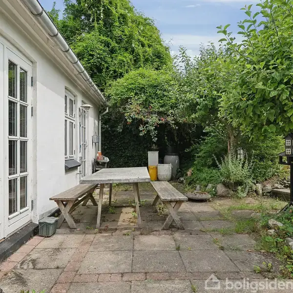 En træbænk og bord står på en flisebelagt terrasse ved et hvidt hus. I baggrunden er der grønne buske og træer, der omgiver området.